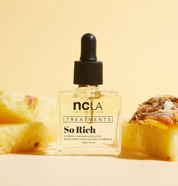 Square glass bottle of NCLA Treatments So Rich cuticle oil with black rubber dropper cap is staged with slices of pineapple