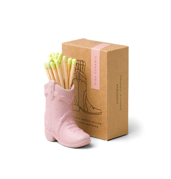 Pink ceramic cowboy boot match holder with green-tipped wooden matches inside next to brown box packaging