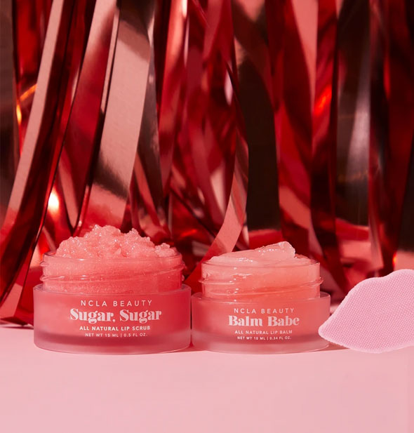 NCLA Beauty Sugar, Sugar and Balm Babe jars with textured lip-shaped scrubber partially shown on a background of red streamers