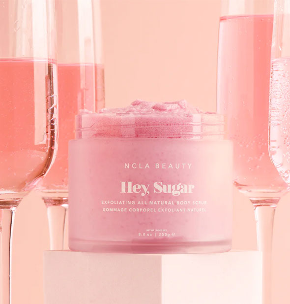 Pot of Pink NCLA Beauty Hey, Sugar Exfoliating All Natural Body Scrub on a rectangular pedestal flanked by flutes of bubbly, pink champagne
