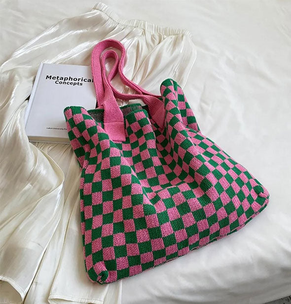 Knit pink and green checkered tote bag with pink straps rests on a white bedspread with a white satin skirt and Metaphorical Concepts book