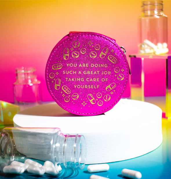 Round glittery pink pill case on platform surrounded by pill bottles says, "You are doing such a great job taking care of yourself" in gold lettering surrounded by gold pill, capsule, and star graphics