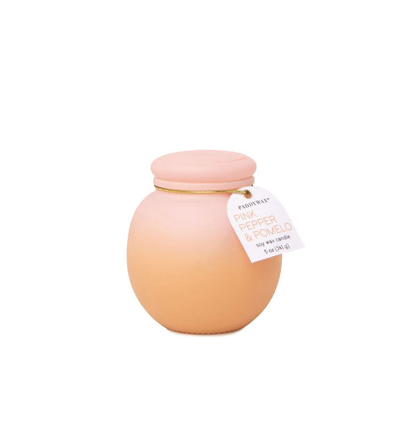 Rounded candle jar with frosted matte finish and orange-to-pink ombre coloration is tpoped with a lid and tag on a gold band