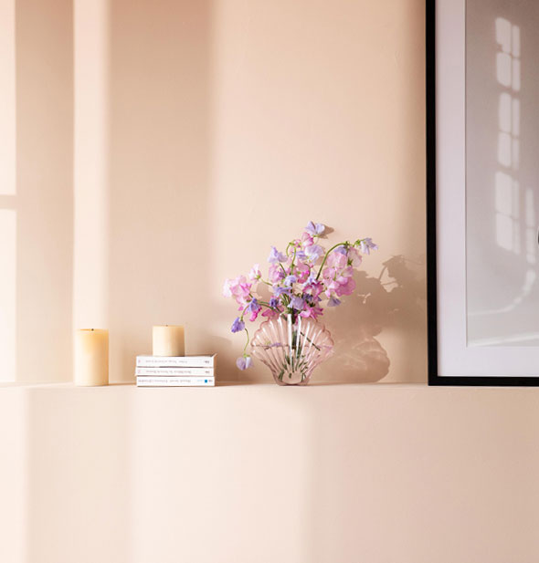 Pink glass seashell vase staged on a shelf with books and pillar candles holds stalks of clustered pink flowers next to a picture frame