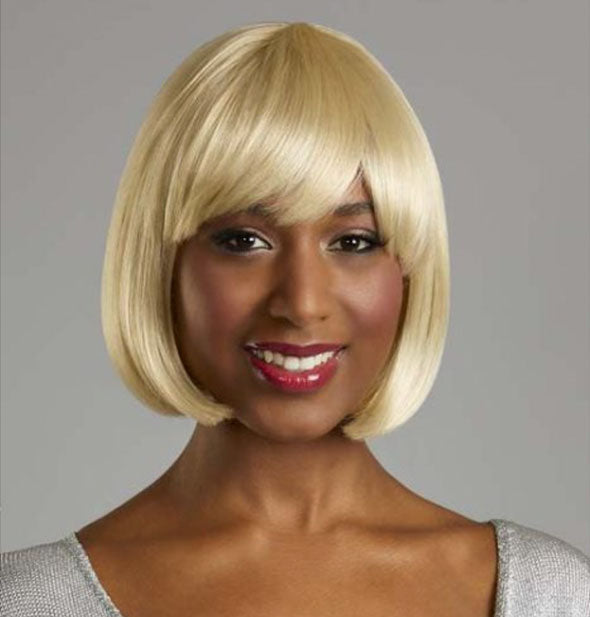 Smiling model wears a short, chin-length, bob-style blonde wig with bang fringe