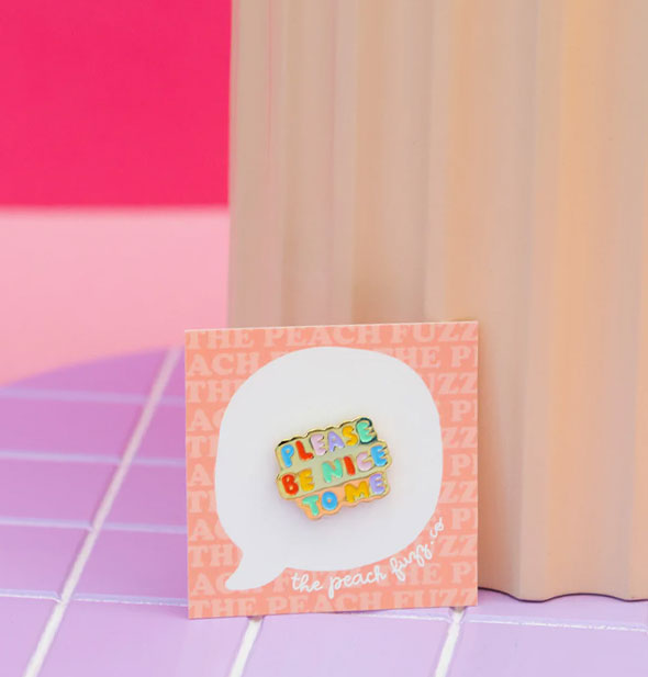 Multicolored enamel Please Be Nice to Me pin on The Peach Fuzz product card is propped against a wavy-edged block on a purple tile surface