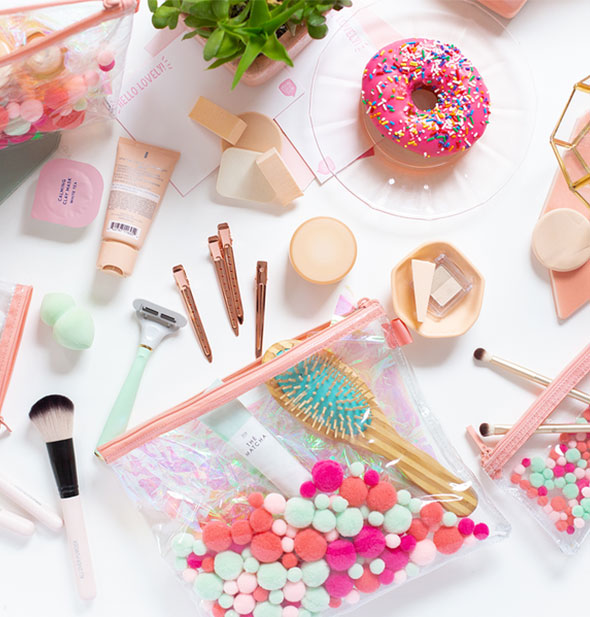 Clear plastic pouch with colorful embedded pom poms on a tabletop with an assortment of toiletries, makeup supplies, and a pink frosted donut with rainbow sprinkles
