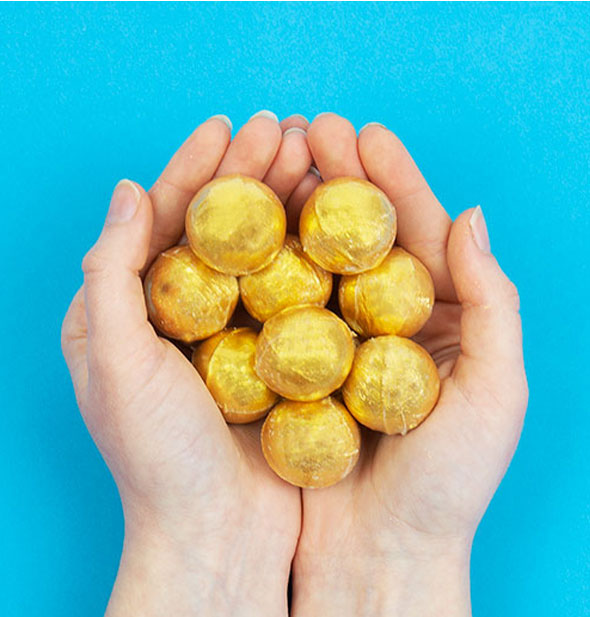 Model's hands cup nine visible round golden bath bombs against a blue background