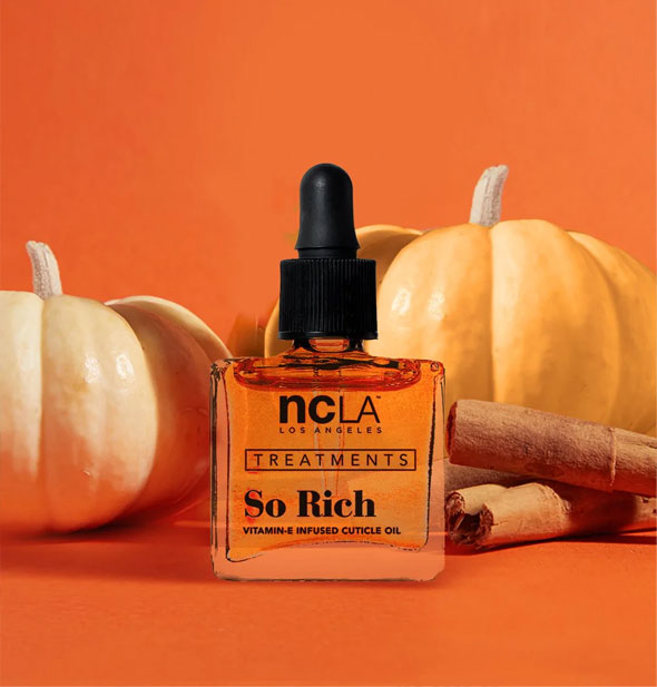 Square glass bottle of NCLA Los Angeles Treatments So Rich Vitamin E-Infused Cuticle Oil with black dropper cap is staged with yellow pumpkins and cinnamon sticks on an orange background