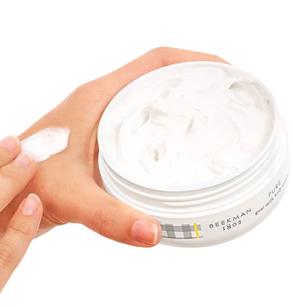Model holding a tub of Beekman 1802 body cream applies a dab of product to back of hand