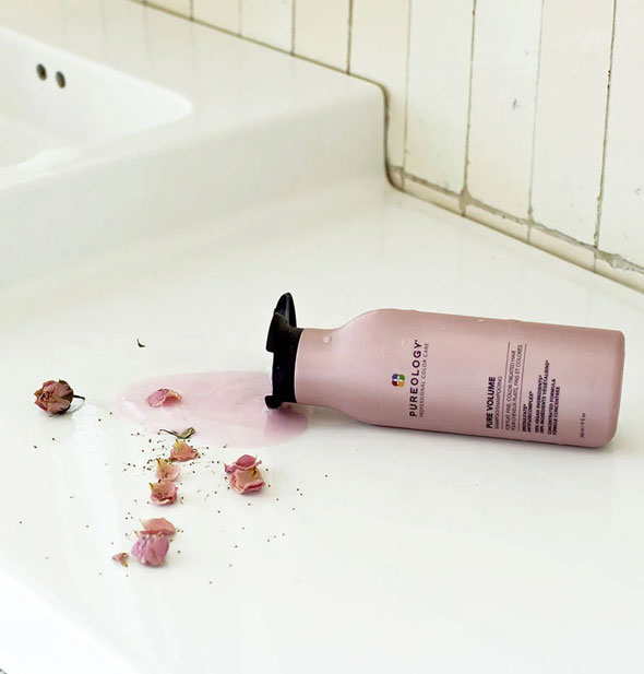 A bottle of Pureology Pure Volume Shampoo lays on its side spilling out some product over dried rose petals