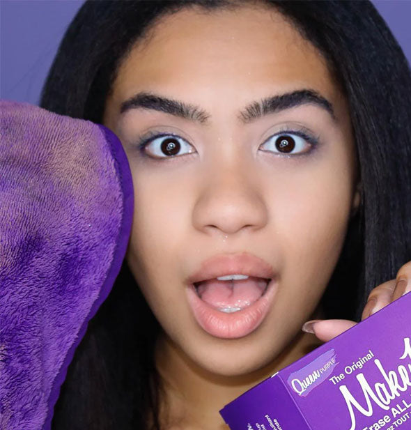 Model with a surprised facial expression holds a Queen Purple Original MakeUp Eraser box in one hand and cloth with makeup smudges on it in the other