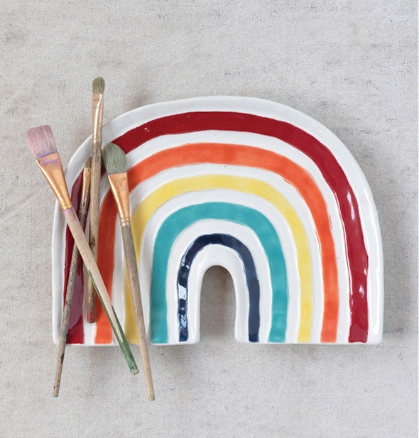 Arced white rainbow platter with painted stripes in red, orange, yellow, teal, and dark blue holds several paint brushes