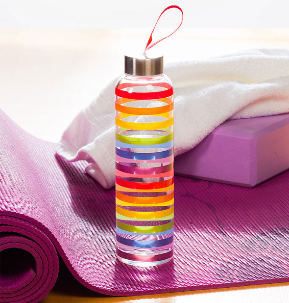 Rainbow striped water bottle sits on a purple yoga mat with towel in background