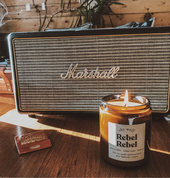 Lit amber glass jar Rebel Rebel candle sits on a wooden tabletop in front of a Marshall amp with a pack of incense matches