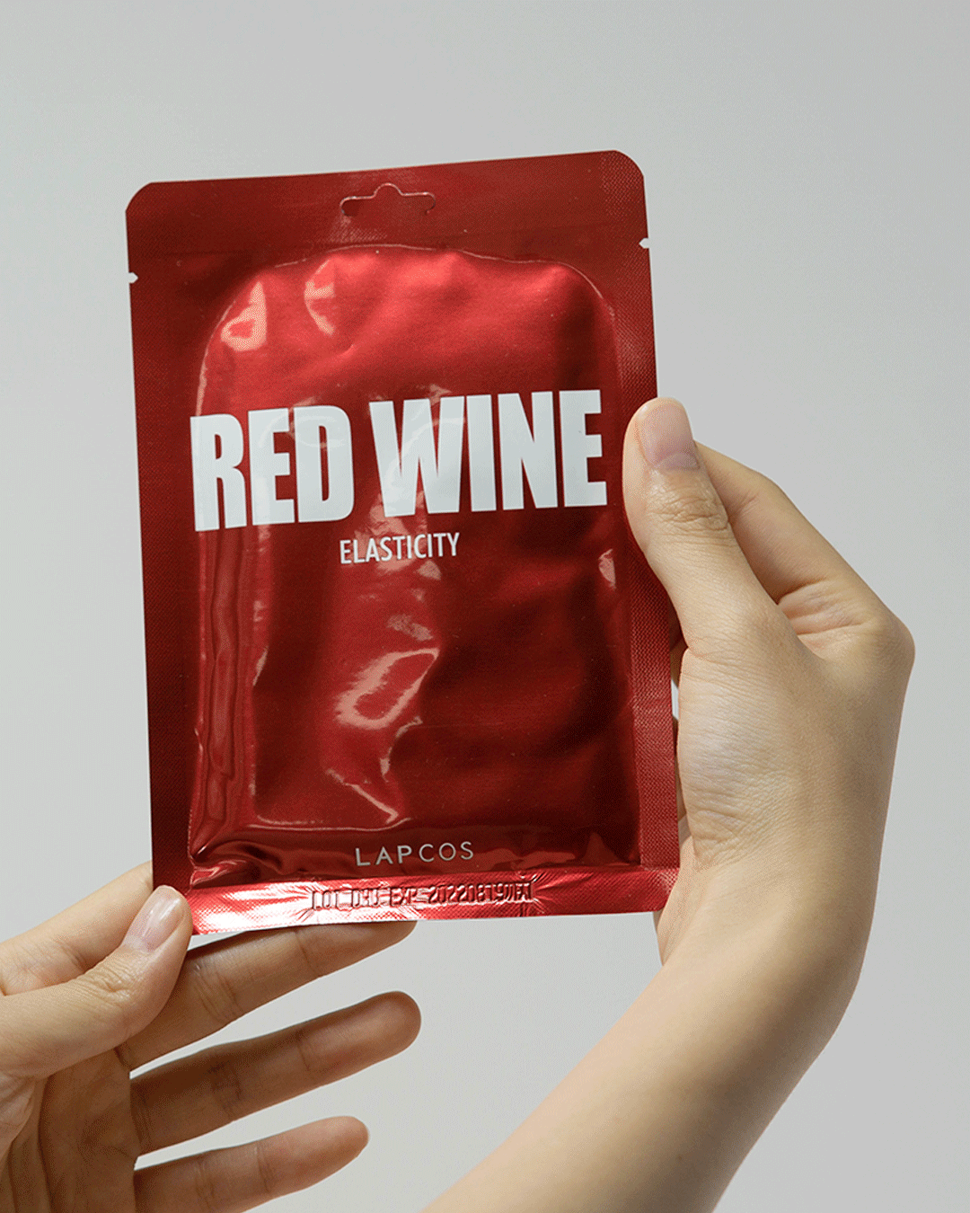 Model's hands open a Red Wine Elasticity sheet mask packet and unfold the mask inside