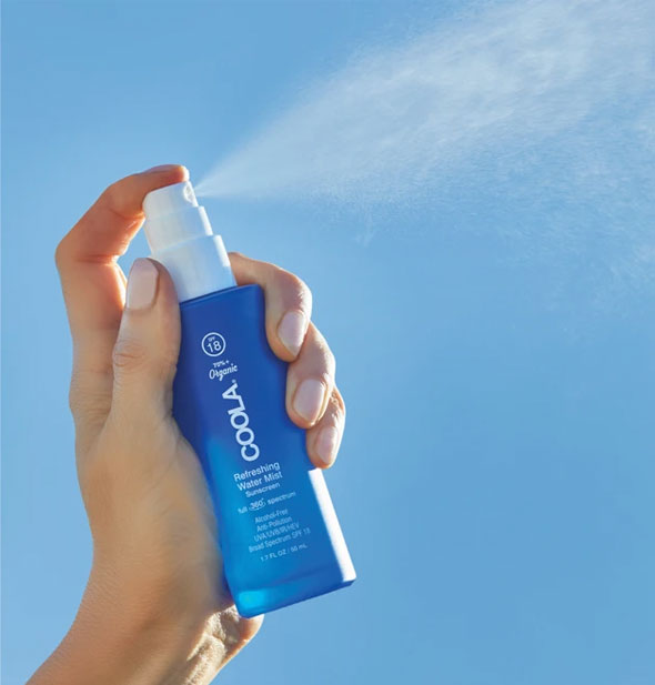 A model's hand holds and sprays from a bottle of COOLA Refreshing Water Mist Sunscreen in front of a blue sky