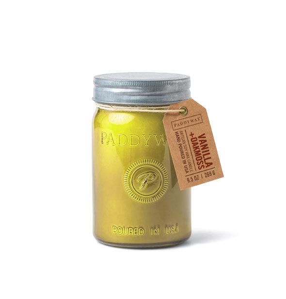 Glass Paddywax candle jar with yellow candle wax, metal lid, and hanging tag that says, "Vanilla + Oakmoss"