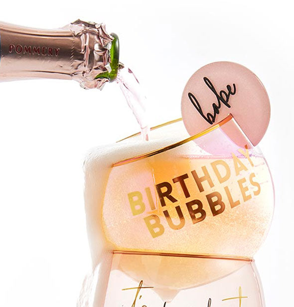 Champagne pours from a bottle into a crookedly-stacked Birthday Bubbles glass with "Babe" tab attached to the rim