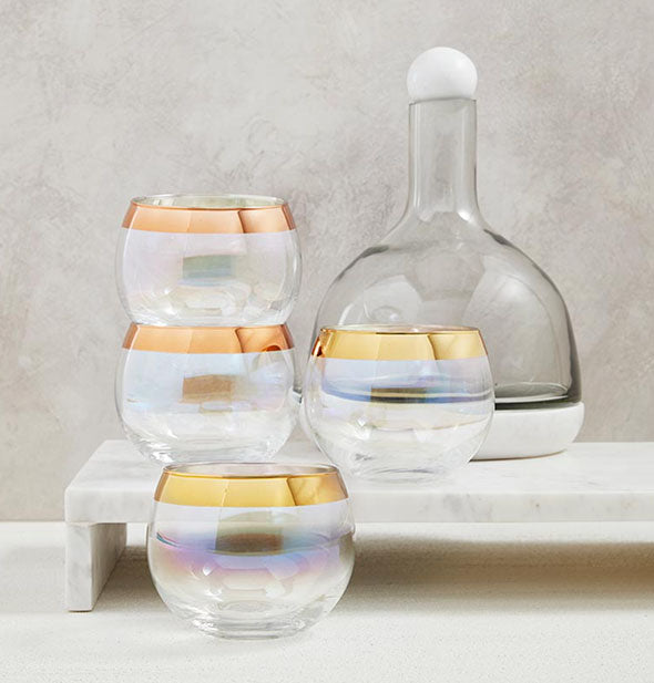Set of four bowl-shaped bar glasses with wide metallic gold rims sit alongside a glass decanter on marble platform
