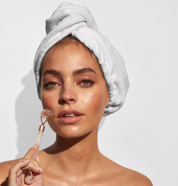 Model wearing hair towel demonstrates use of a gemstone facial roller on cheek