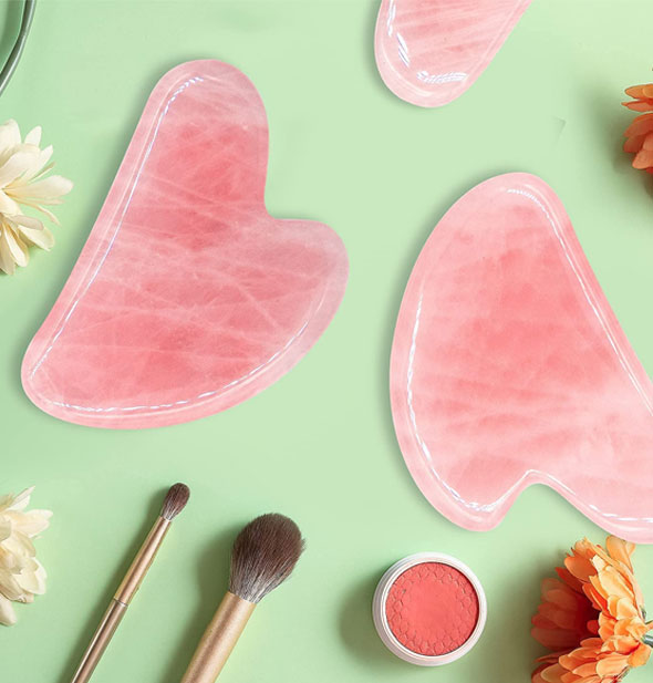 Rose quartz gua sha massagers on a green surface staged with flowers, makeup brushes, and a pink cosmetic