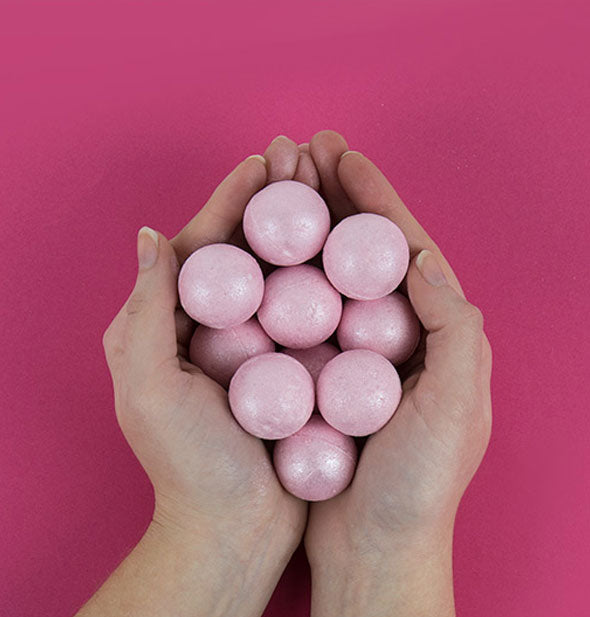 Model's hands cup a handful of pearly pink bath bombs against a magenta backdrop