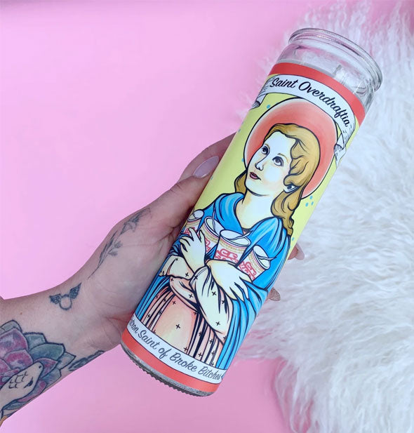 A hand holds the Saint Overdraftia, Patron Saint of Broke Bitches prayer candle against a pink and white background