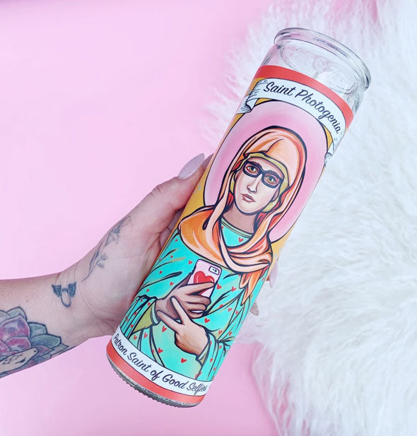 A hand holds the Saint Photogenia, Patron Saint of Good Selfies illustrated prayer candle against a pink and white fur background