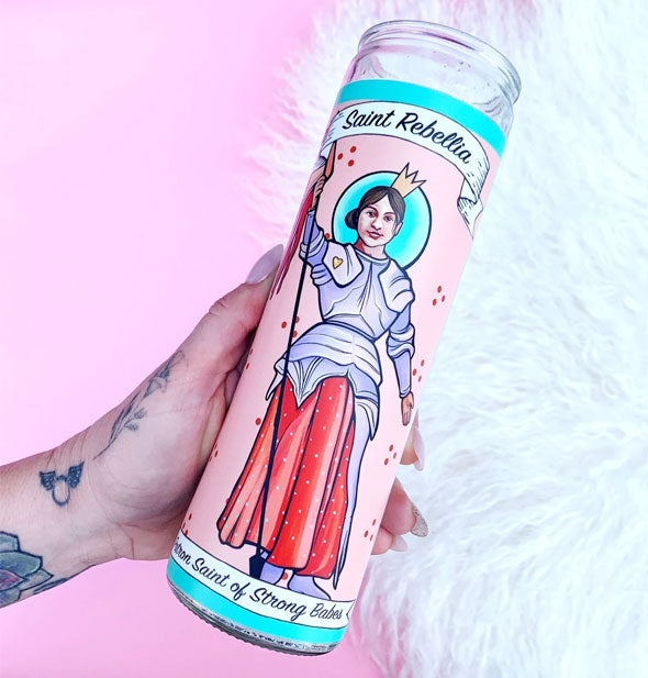 A hand holds the Saint Rebellia, Patron Saint of Strong Babes prayer candle against a pink and white fur background