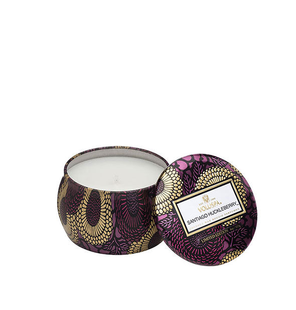 A small unlit candle inside a rounded tin with metallic purple and gold floral design and matching lid set to the side.
