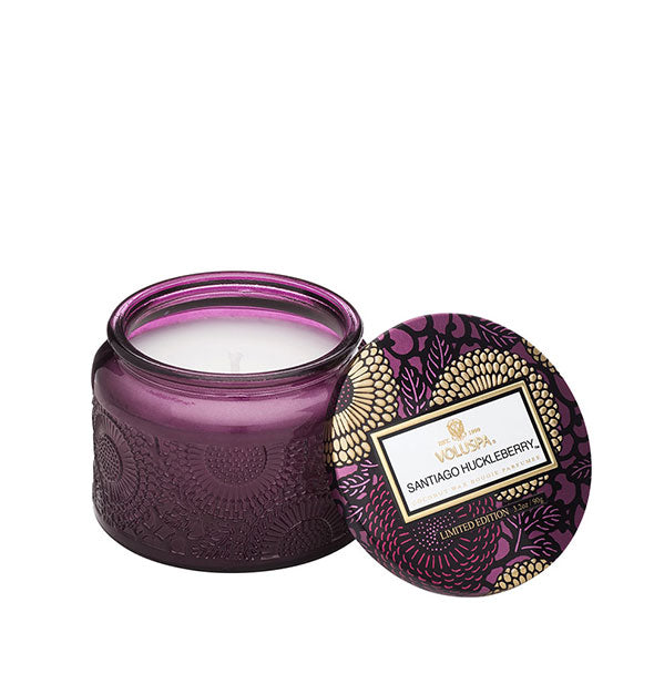 Embossed purple glass Santiago Huckleberry Voluspa candle jar with purple and gold metallic floral lid set to the side