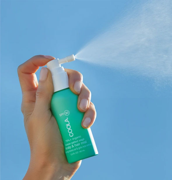 A model holds and sprays from a bottle of COOLA Scalp & Hair Mist in front of a blue sky