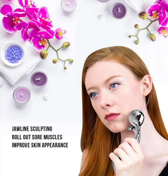 Model uses Sculpting Massager on neck underneath accents of purple flowers and candles with caption, "Jawline sculpting; Roll out sore muscles; Improve skin appearance"