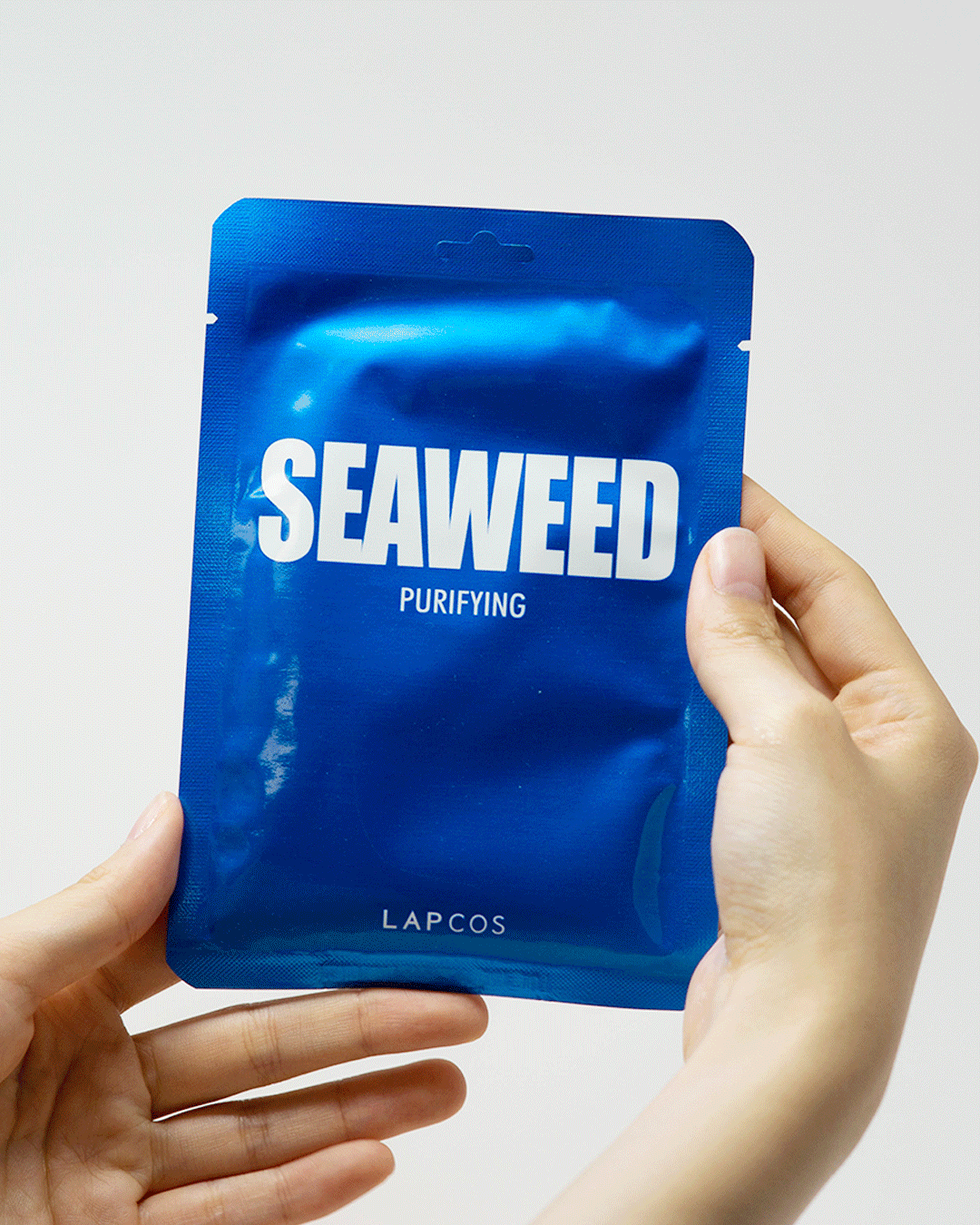 Model's hands open a Seaweed sheet mask pack and unfold the mask inside