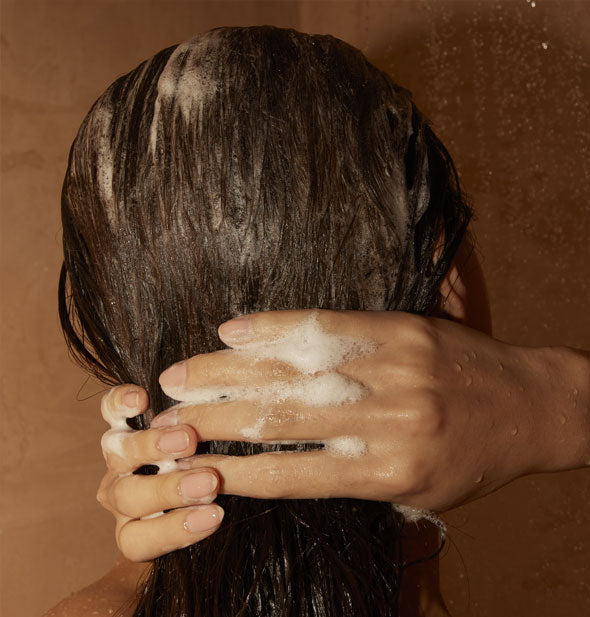 Model wrings shampoo lather from wet hair under running shower water