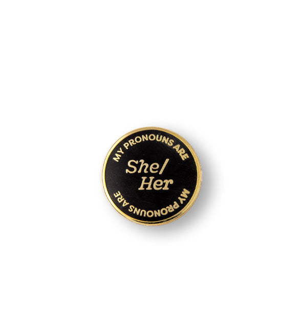 Round black enamel pin with gold border says, "My Pronouns Are" at top and bottom and "She/Her" in the center, all in gold lettering