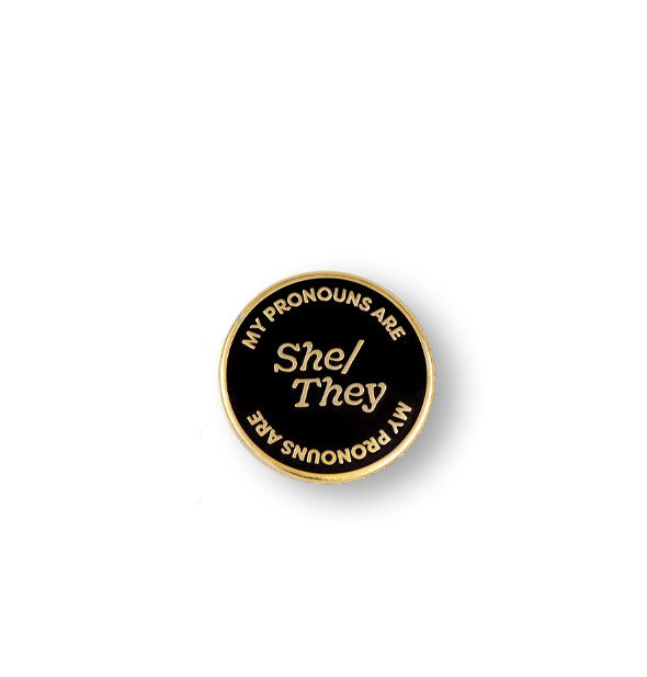 Round black enamel pin with gold border says, "My Pronouns Are" at top and bottom and "She/They" in the center, all in gold lettering