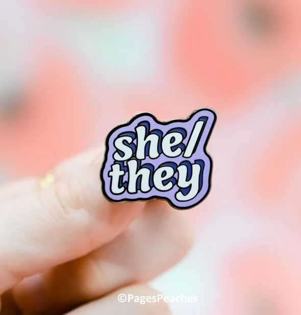 A purple enamel She/They pin is held between a model's fingers with Pages Peaches image copyright at bottom