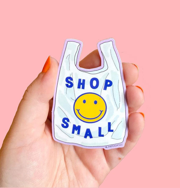 Model's hand holds a shopping bag-shaped sticker that says, "Shop Small" with smiley face graphic