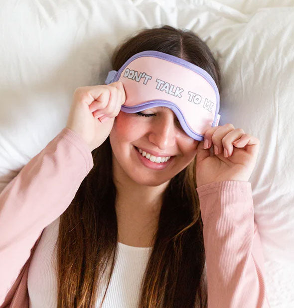 Smiling model laying on white linens pulls a Don't Talk to Me sleep mask over eyes