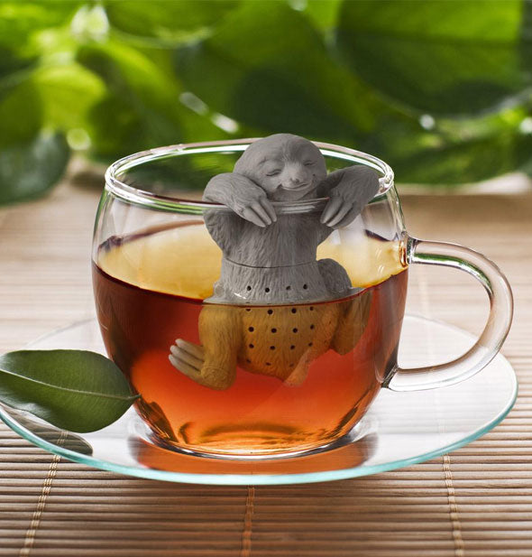 Sloth tea diffuser rests on the edge of a clear glass mug with brewed tea inside