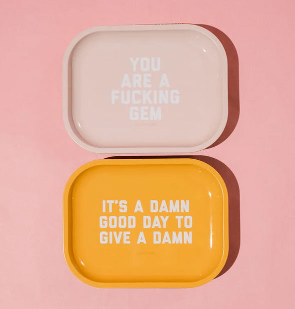 Two rectangular trays with rounded corners, one light pink and the other bright yellow; the top pink tray says, "You are a fucking gem" in light lettering and the bottom yellow tray says, "It's a damn good day to give a damn" in white lettering