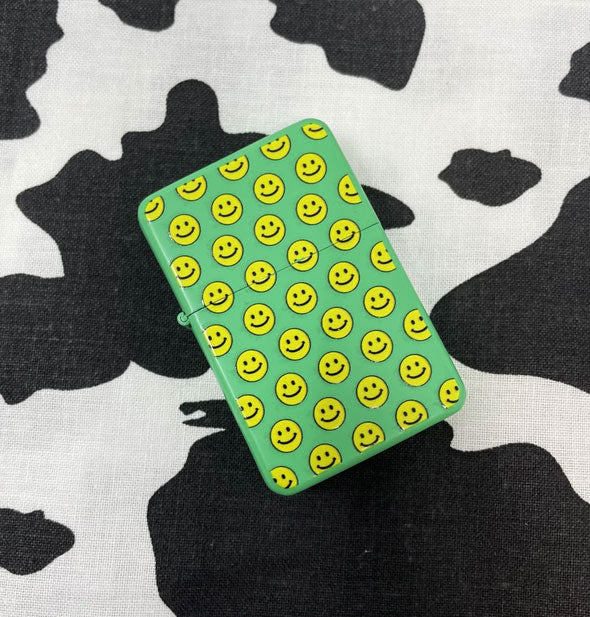 Green lighter with all-over yellow smiley face print sits on a cowhide print backdrop