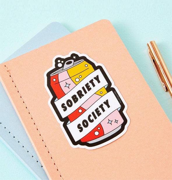Sticker on a peach-colored notebook says, "Sobriety Society" in a white banner wrapped around a soda can with fizz and star graphics