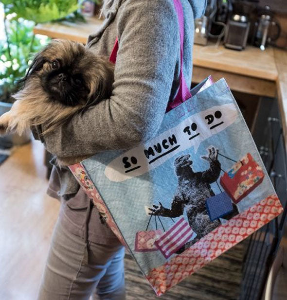 Model cradling a brown Shih Tzu poses with the So Much to Do Godzilla shopper hanging from her shoulder 