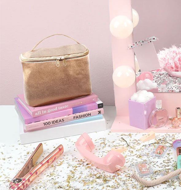 Gold makeup bag is staged on a vanity with books, telephone, perfume, cotton balls, flat iron, and other items