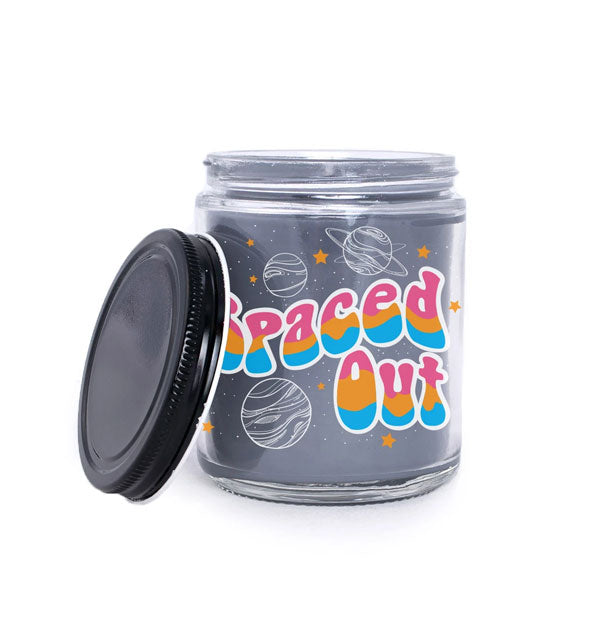 Black wax candle in clear glass jar with black lid says, "Spaced Out" in rainbow font surrounded by planets and stars