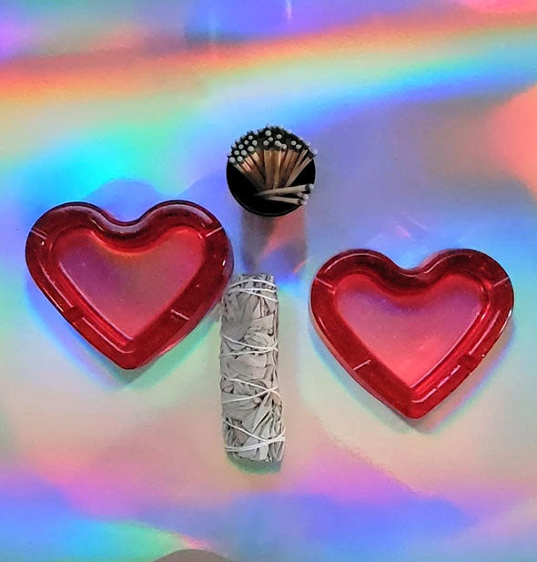 Two red heart-shaped ashtrays staged with matches and a sage bundle on holographic surface