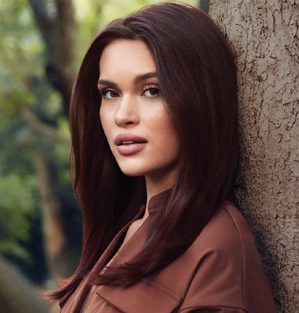Model with healthy-looking straight brown hair leans against a tree